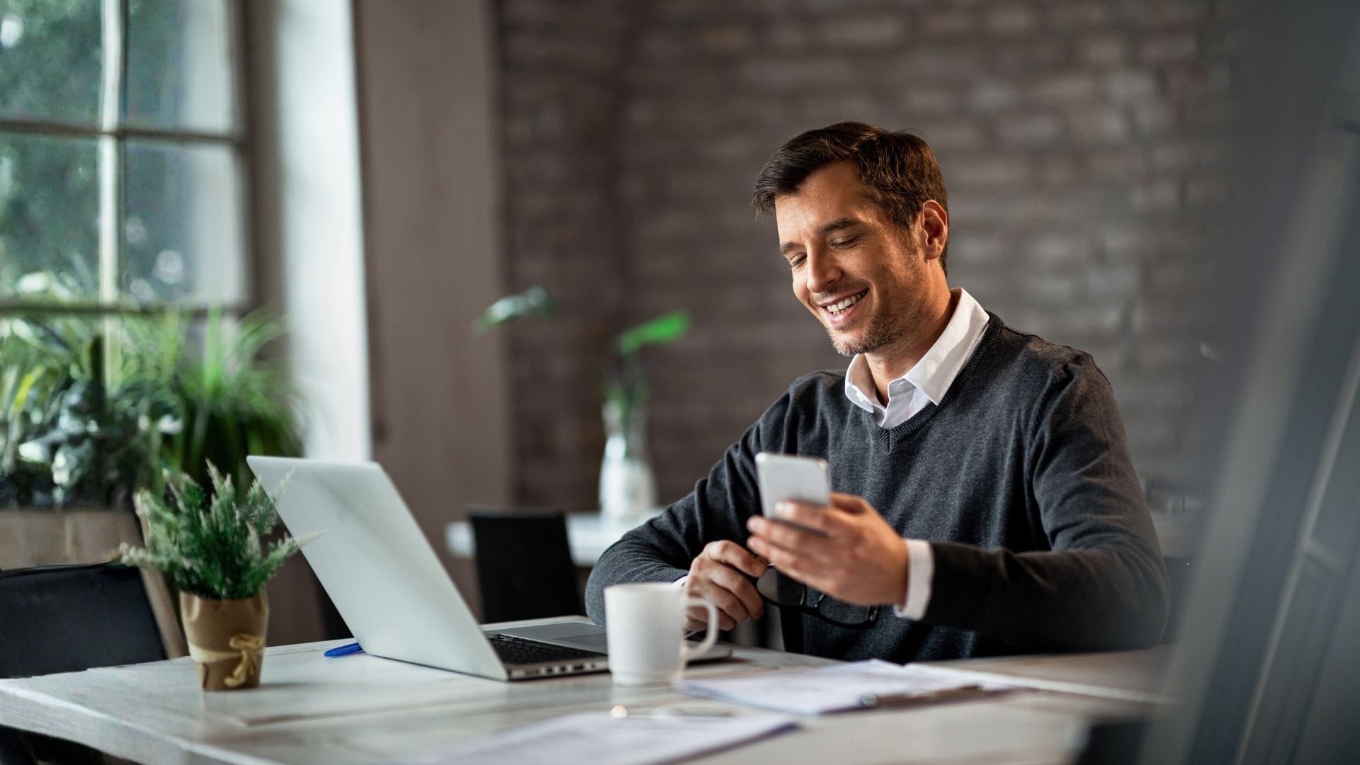 Executive at a desk with laptop and coffee reviewing charter flight booking details on a smartphone.